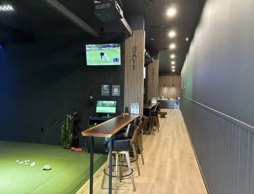 An indoor golf simulator area with a green mat, golf balls, golf clubs, bar-height tables and chairs, and TVs displaying golf. The space has dark walls and wood flooring, leading to a counter in the back.