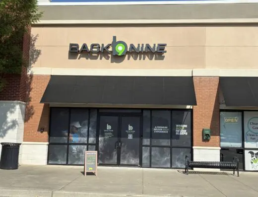Storefront of Back 9 Nine with black awning, large sign, glass doors, sidewalk chalkboard, nearby bench, trash can, and an open sign to the right. Sunlight creates shadows on the sidewalk.
