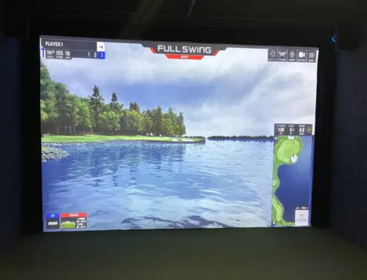 A virtual golf game is displayed on a large screen, showing a golf course with water, trees, and a blue sky. Player stats and course information are visible on the screen.