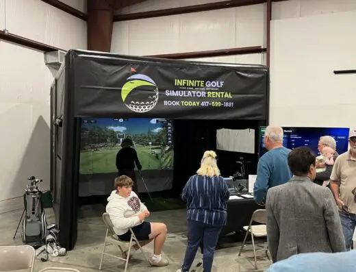 People gather around an indoor golf simulator booth labeled INFINITE GOLF SIMULATOR RENTAL in a large room. Some are seated, while others stand near the entrance or watch the screen inside.