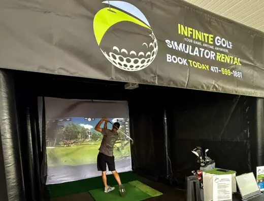 A person swings a golf club in front of a screen at an indoor golf simulator rental, with a large Infinite Golf Simulator Rental banner overhead and golf equipment nearby.
