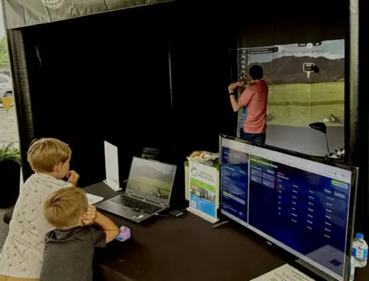 Two boys watch golf simulator screens at an event booth. A man plays golf in the simulator area behind them. The booth sign advertises INFINITE GOLF Simulator Rental. Various screens and promotional materials are on the table.