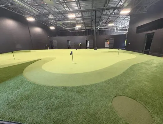 Indoor mini-golf course with artificial grass, multiple putting holes, black walls, and ceiling with exposed pipes and lights. The area appears spacious and well-lit.