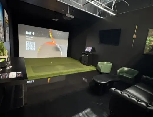Modern indoor golf simulator room with a large screen, artificial turf, lounge seating, a TV, a sword on the wall, and glossy black floors under an open ceiling with exposed pipes and wires.