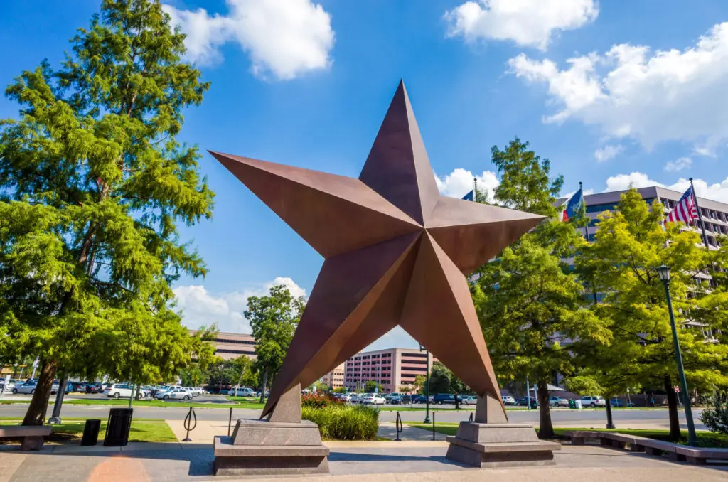 A large bronze star sculpture stands outdoors, surrounded by trees and buildings under a blue sky with clouds.