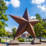 A large bronze star sculpture stands outdoors, surrounded by trees and buildings under a blue sky with clouds.