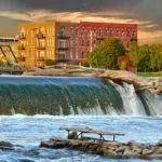 A large fallen tree rests atop a small waterfall with rushing water, while a brick apartment building and green trees stand in the background under a colorful sky. A picnic table sits on rocks in the foreground.