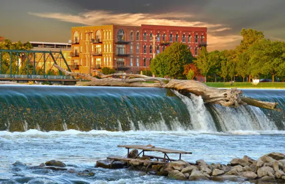 A large fallen tree rests atop a small waterfall with rushing water, while a brick apartment building and green trees stand in the background under a colorful sky. A picnic table sits on rocks in the foreground.