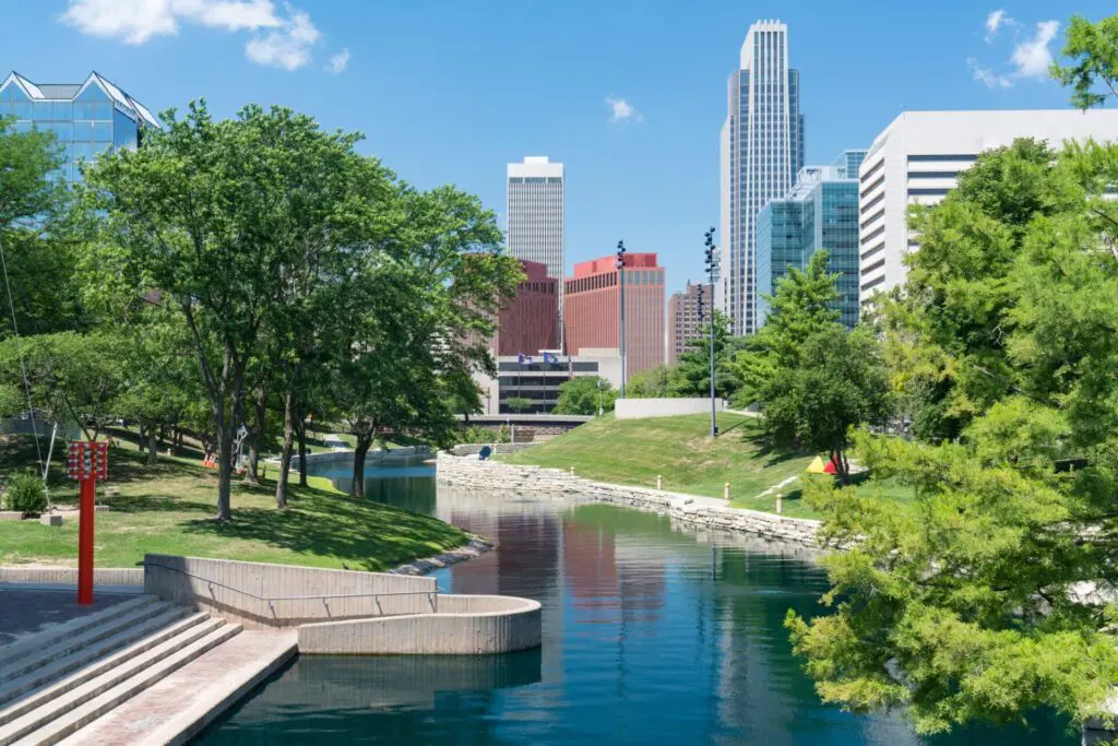 A calm canal lined with trees runs through a city park, with modern high-rise buildings and a clear blue sky in the background on a sunny day.