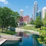 A calm canal lined with trees runs through a city park, with modern high-rise buildings and a clear blue sky in the background on a sunny day.