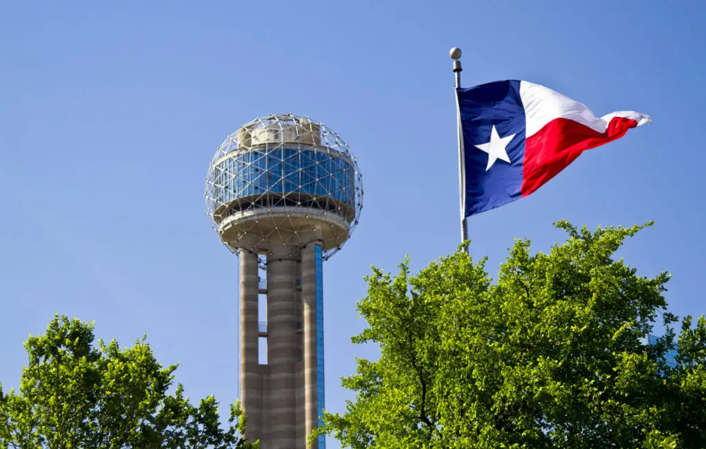 A Texas flag waves near the Reunion Tower in Dallas, with a clear blue sky and green trees in the foreground.