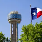 A Texas flag waves near the Reunion Tower in Dallas, with a clear blue sky and green trees in the foreground.