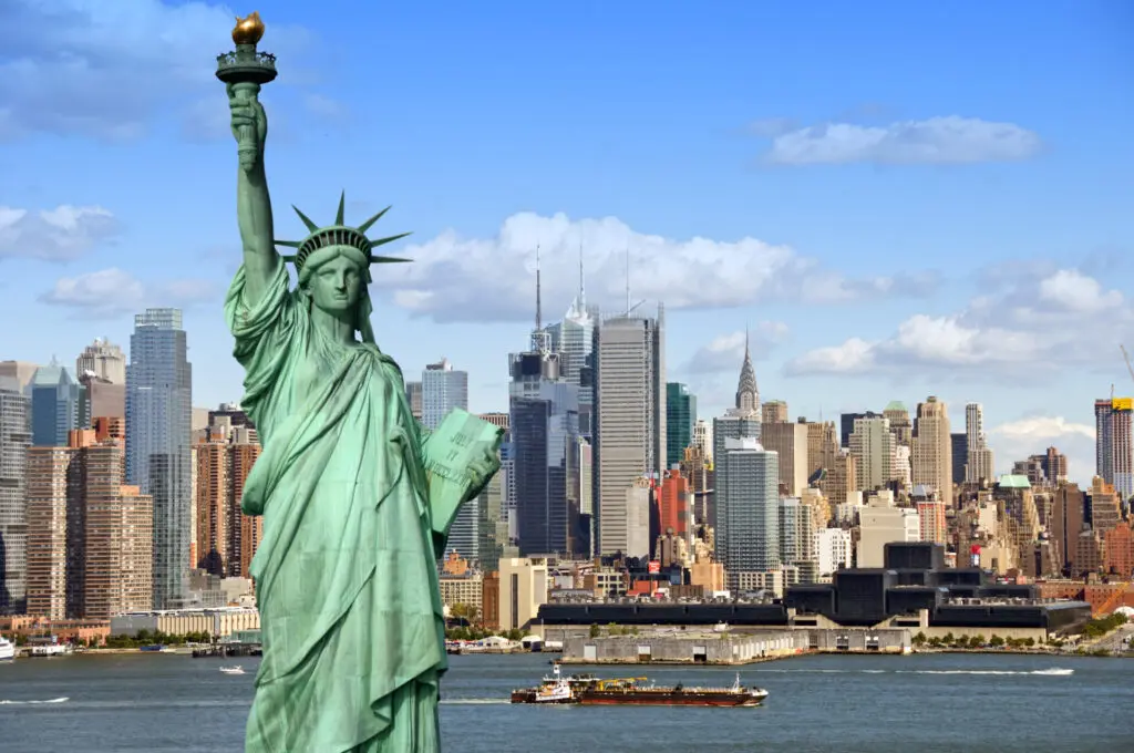 The Statue of Liberty stands in the foreground with the New York City skyline and a boat on the water in the background under a blue sky with clouds.