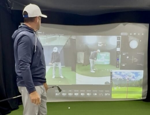 A man in a cap and hoodie holds a golf club, studying a large screen with swing analysis, videos, data, and graphics in an indoor practice area with artificial turf featuring a golf simulator.