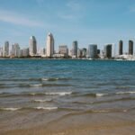 San Diego skyline with modern skyscrapers seen from across the bay on a clear day, gentle waves in the foreground, and blue sky above—a perfect backdrop for a round on a golf simulator or an afternoon stroll.