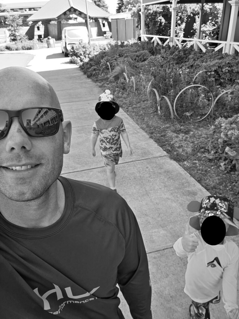 A man wearing sunglasses and a sports shirt takes a selfie while two young children walk behind him on a sidewalk near greenery and buildings. The children wear sun hats and casual clothes. The image is in black and white.