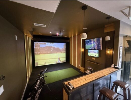 A modern indoor golf simulator room with a large screen displaying a virtual golf course, golf clubs on the floor, two wall-mounted TVs, bar seating with stools, and a wooden counter under warm lighting.