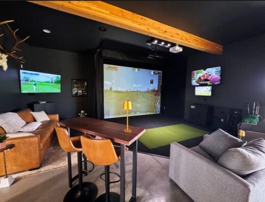 A modern golf simulator room with a large screen, two TVs on the wall, a brown leather couch, a gray sofa, bar stools at a tall table, and golf green mat on the floor. Exposed wooden beam and decor visible.