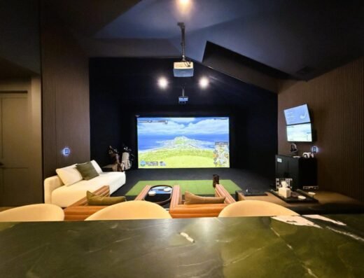 A modern golf simulator room with a large screen displaying a golf course, cozy seating including sofas and armchairs, a green putting area, wall-mounted TV, and golf clubs in the corner.