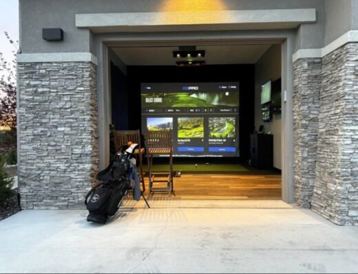 A home garage converted into a golf simulator, with a golf bag and clubs in the foreground and a large screen displaying golf graphics on the back wall. Two chairs face the screen, and the garage has stone accents.