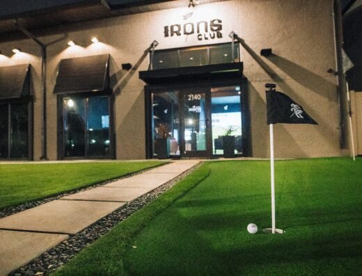 A putting green with a golf ball and flag in the foreground leads up to the entrance of a building labeled IRONS CLUB at night. The walkway is lined with stones and the area is well-lit.