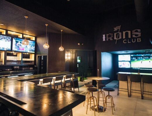 Modern sports bar interior with a long bar counter, barstools, and several flat-screen TVs displaying sports. The wall features the IRONS CLUB logo. The space is dimly lit with hanging pendant lights.