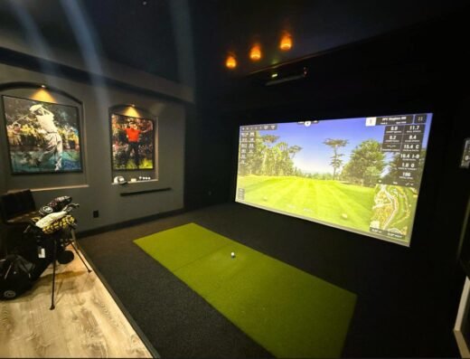 A home golf simulator room with a large screen showing a golf course, a green mat with a golf ball, golf clubs on the left, wood flooring, and framed golf-themed artwork on the wall.