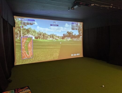 A golf simulator room with a large screen displaying a golf game, green turf flooring, a golf ball on the floor, and part of a sign visible in the lower left corner.