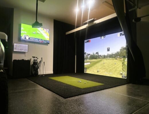 A home golf simulator setup with a large projector screen displaying a golf course, artificial turf mat, golf clubs nearby, and a TV on the wall showing a football game. Bright overhead lights illuminate the space.