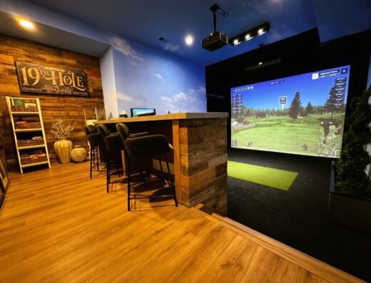 A modern indoor golf simulator room with a large screen showing a virtual golf course, green turf mat, bar seating, and wood-paneled walls with a 19th Hole sign. The atmosphere is cozy and inviting.