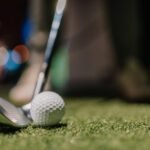 A close-up of a golf club and golf ball on green artificial turf, ready for a shot, with blurred lights and a golfer’s shoes visible in the background.