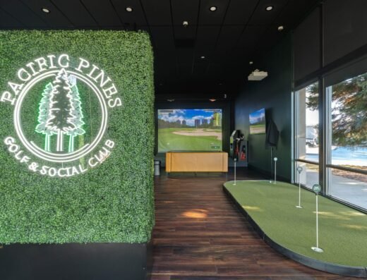Indoor golf facility with a putting green, golf simulator screen, and a wall covered in greenery featuring a neon sign that reads Pacific Pines Golf & Social Club. Large windows provide natural light.