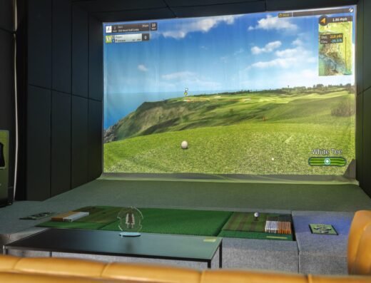A golf simulator room with a large screen displaying a virtual golf course, golf mats on the floor, and golfing equipment nearby. A golf ball is positioned on the mat in front of the screen.