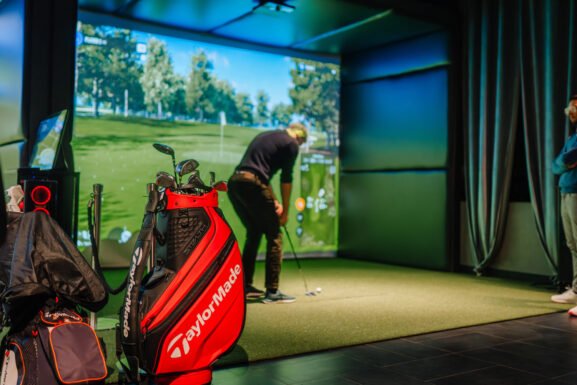 Golfer lining up a putt inside a golf simulator bay at an indoor golf venue, with a TaylorMade staff bag in the foreground